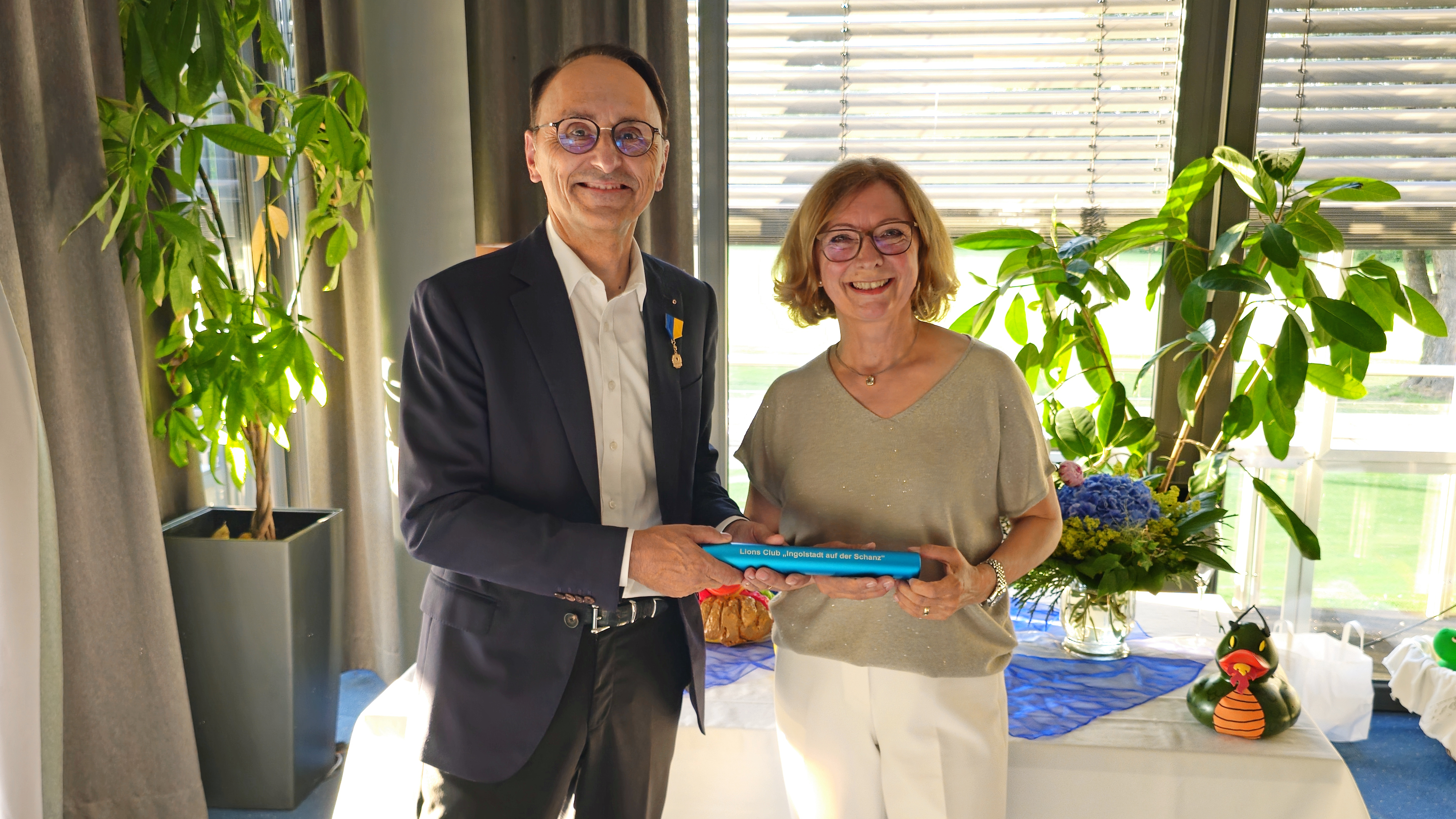Maria Garnisch und Richard Riedmaier halten gemeinsam den blauen Staffelstab mit der Aufschrift „Lions Club Ingolstadt auf der Schanz“ in die Kamera.