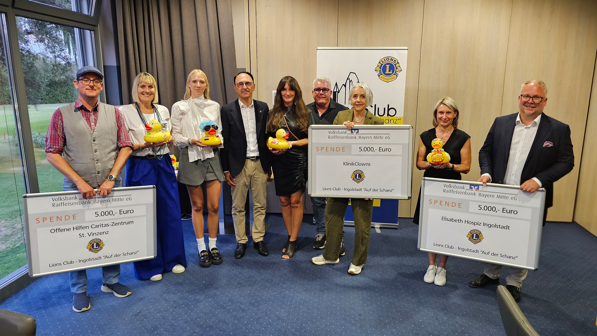 Gruppenfoto: Vertreterinnen und Vertreter der drei Einrichtungen halten gemeinsam mit Mitgliedern des Lions Club Ingolstadt „Auf der Schanz“ drei Spendenschecks über je 5.000 Euro und gelbe Enten in die Kamera.